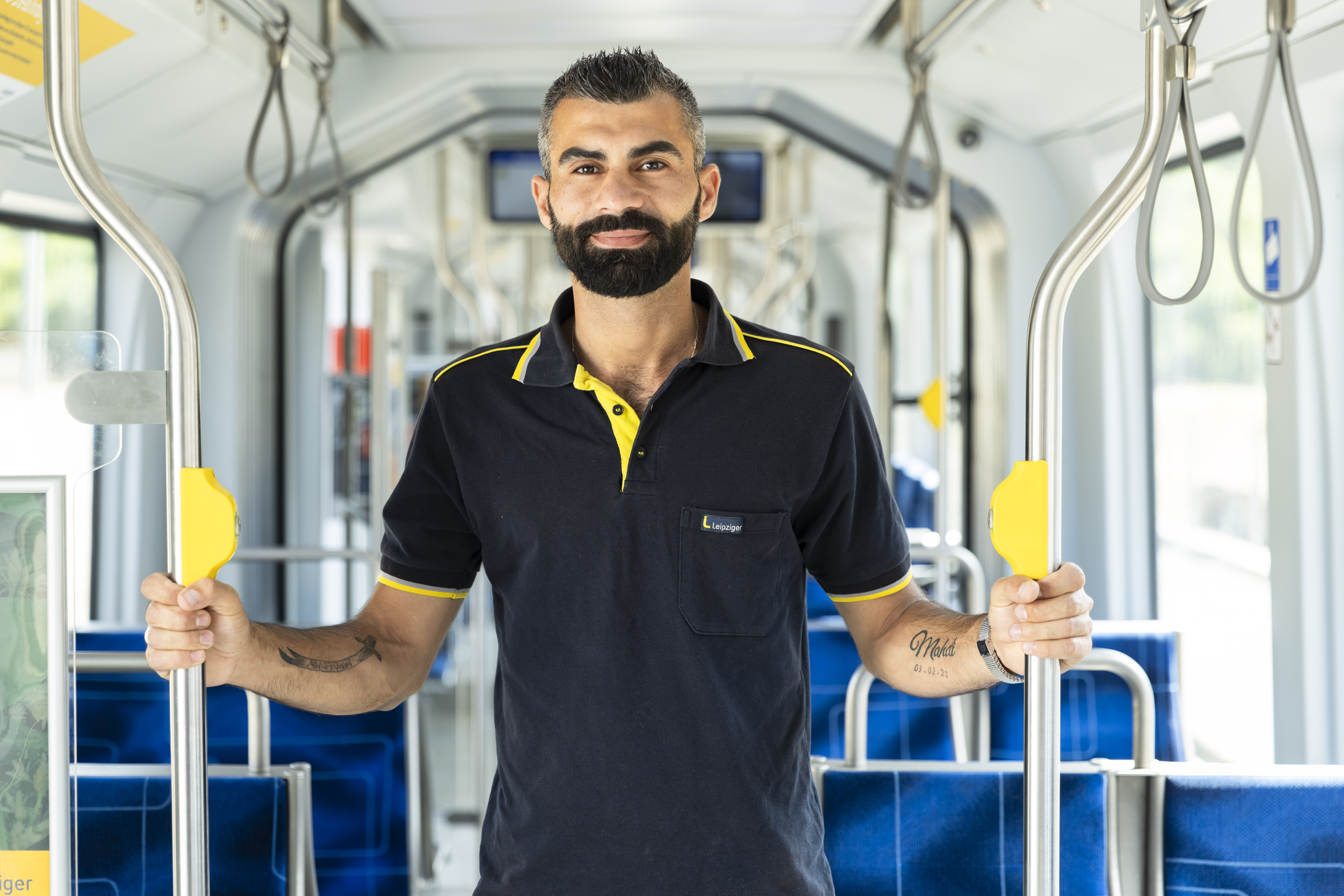 Nazar Arab steht in einer leeren Straßenbahn. Er hält sich mit beiden Händen an den Metallstäben fest und lächelt in die Kamera.